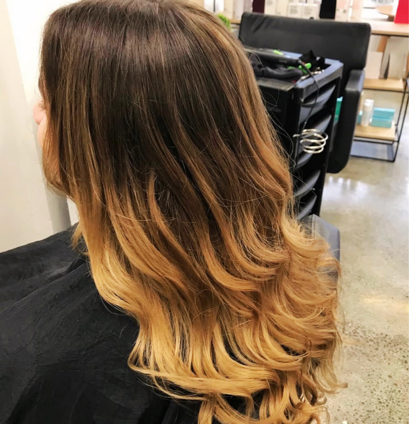 A person with shoulder-length hair styled with dark roots and blonde, wavy ends, sitting in a salon chair.