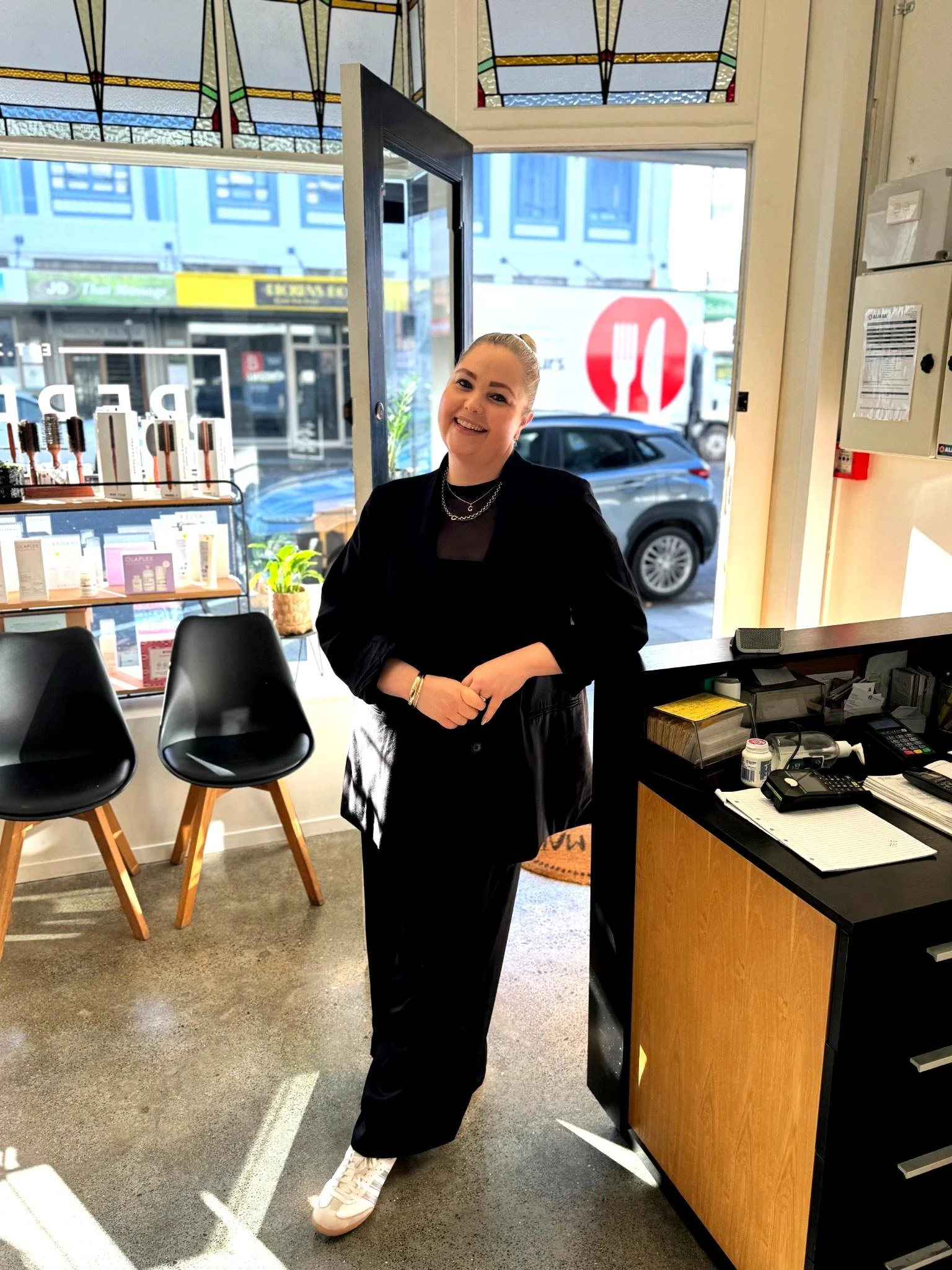 A woman stands inside a cafe by the entrance, smiling at the camera. She is wearing a black outfit and white sneakers, with hair pulled back. The cafe has a counter with office supplies and a few black chairs near a window showing a street scene outside.