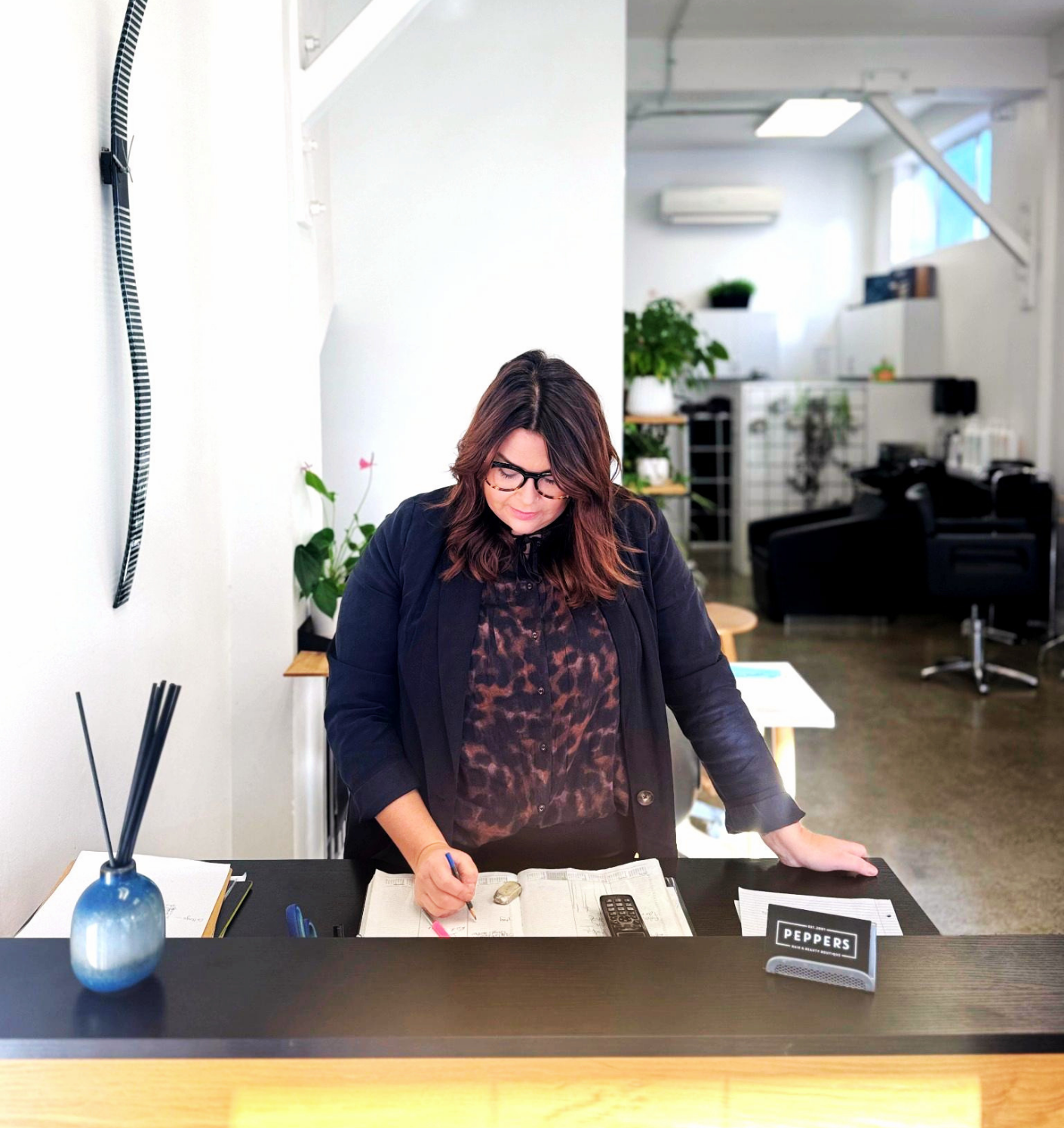 A woman with brown hair, black glasses, and a dark blazer is working at a reception desk. She is writing on a piece of paper with a highlighted marker. There is a blue vase with black reed diffusers, a remote control, and some documents on the desk. The background shows a modern, well-lit office or reception area with green plants and seating.