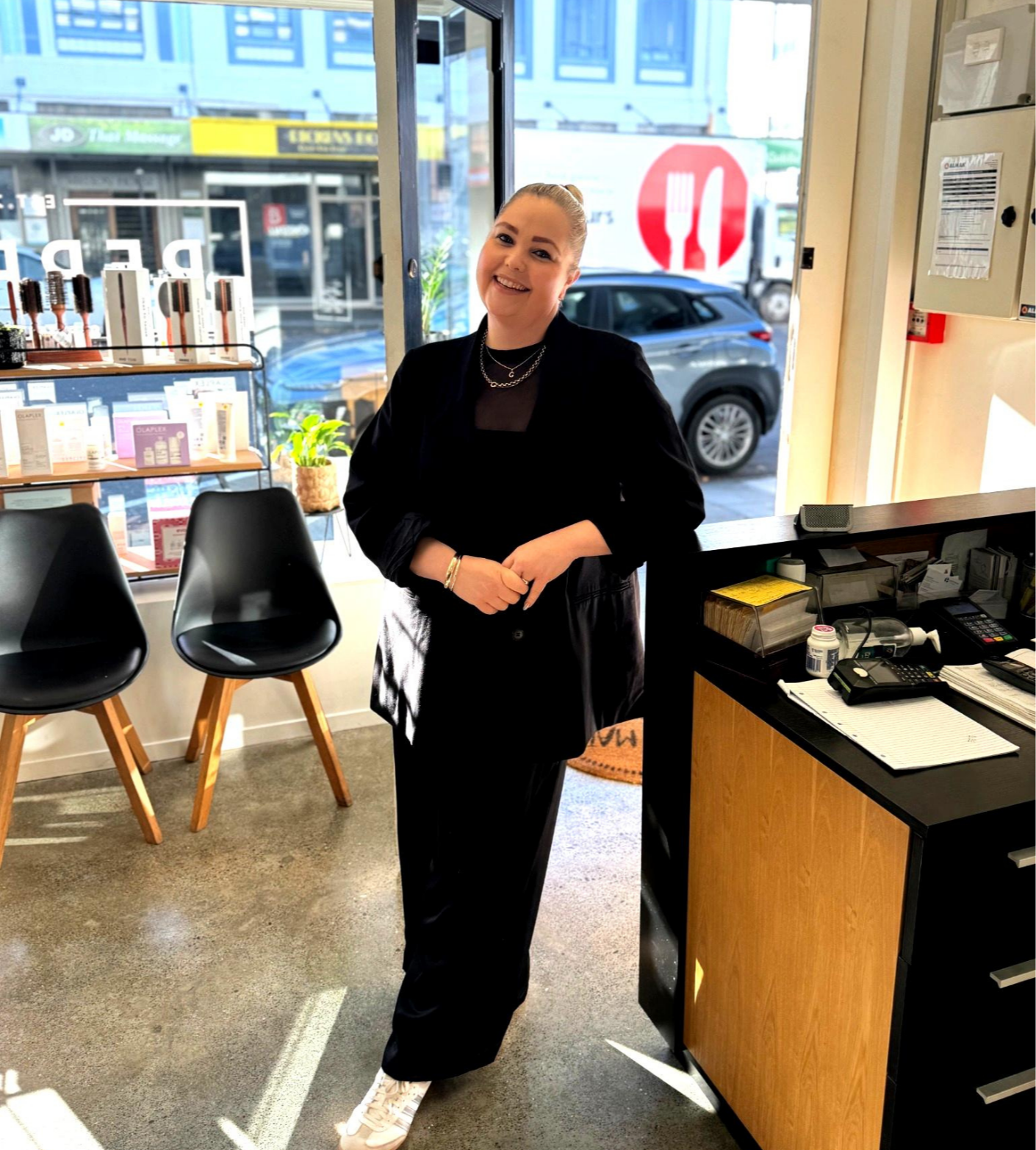 A woman standing inside a cafe or restaurant, smiling, dressed in black, with chairs and display shelves behind her, and a window showing a street scene outside.