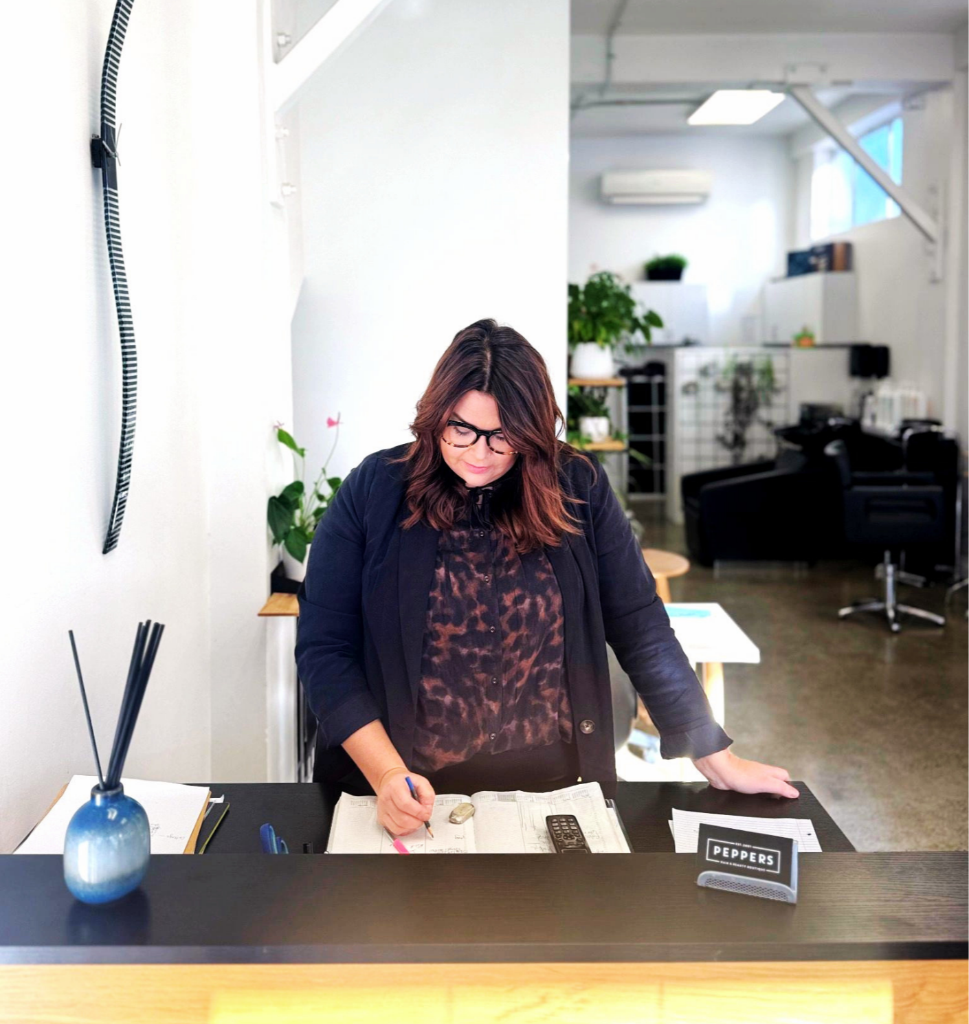 A woman with dark hair, glasses, and a black blazer stands at a reception desk, writing on a paper with a pen. The desk has a blue vase with black sticks, a calculator, and a sign reading 'PEPPERS'. The background shows a modern, well-lit office space with plants and black lounge chairs.