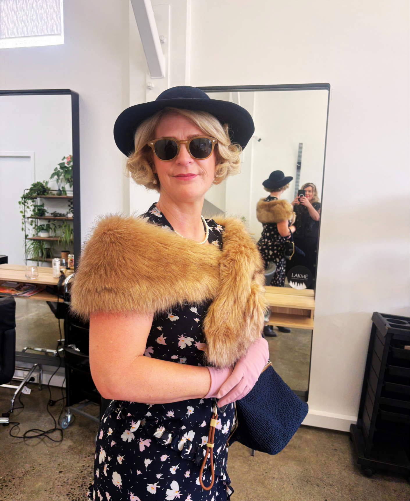 A woman dressed in a floral dress, accessorized with a fur stole, pink gloves, sunglasses, a black hat, and pearls, standing in front of a mirror at a salon or studio.