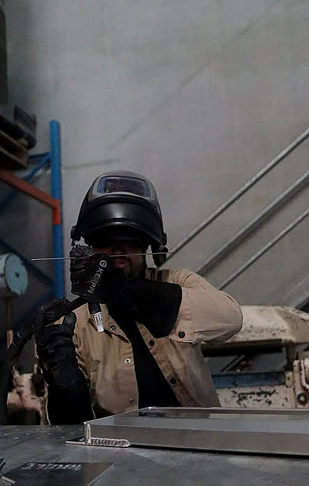 A person wearing protective welding gear, including a helmet and gloves, is welding a metal piece in an industrial workshop.