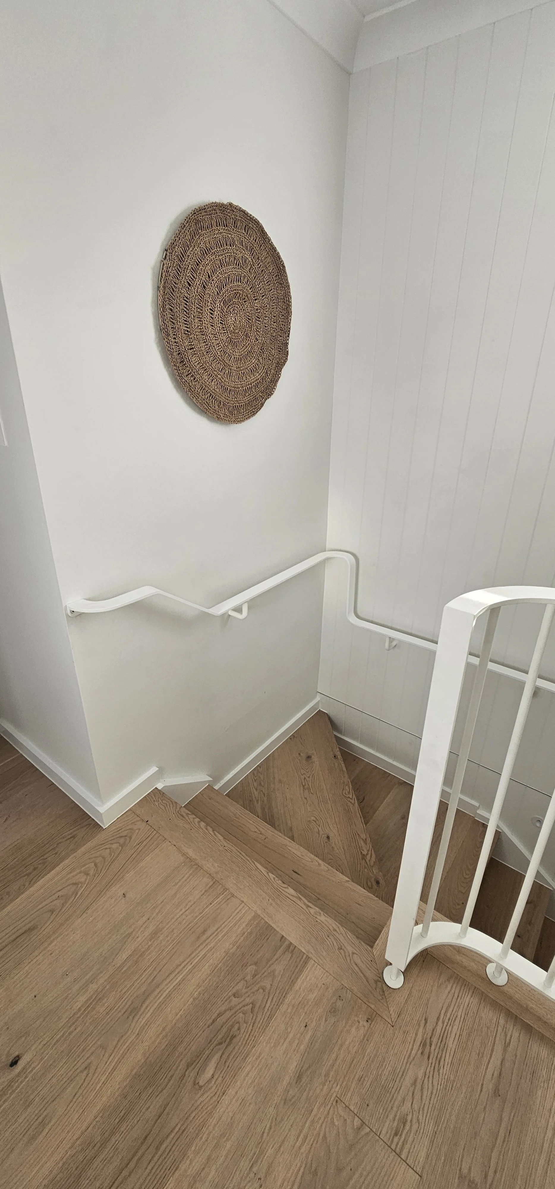 Wooden staircase descending in a white-walled space with a woven round wall decoration and a white handrail.