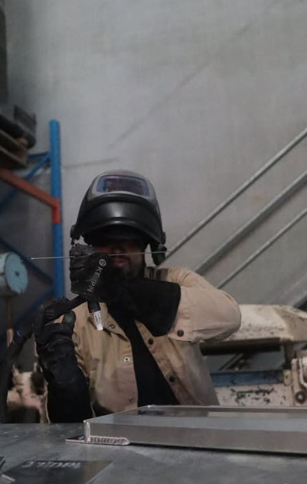 Person wearing a welding helmet and gloves, holding a welding tool in an industrial workshop.