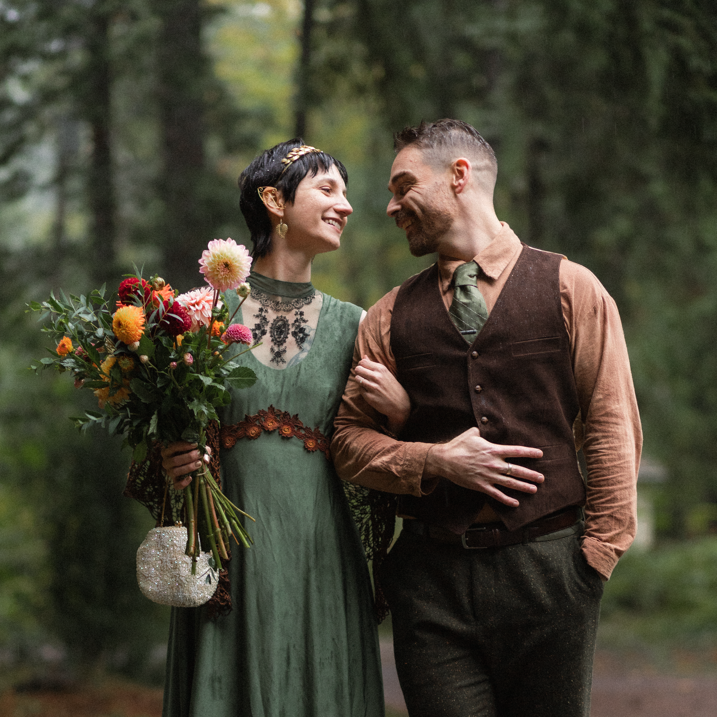 A smiling woman holding a bouquet of colorful flowers and a small beaded purse, standing in an outdoor forest setting with a man, both facing each other affectionately.