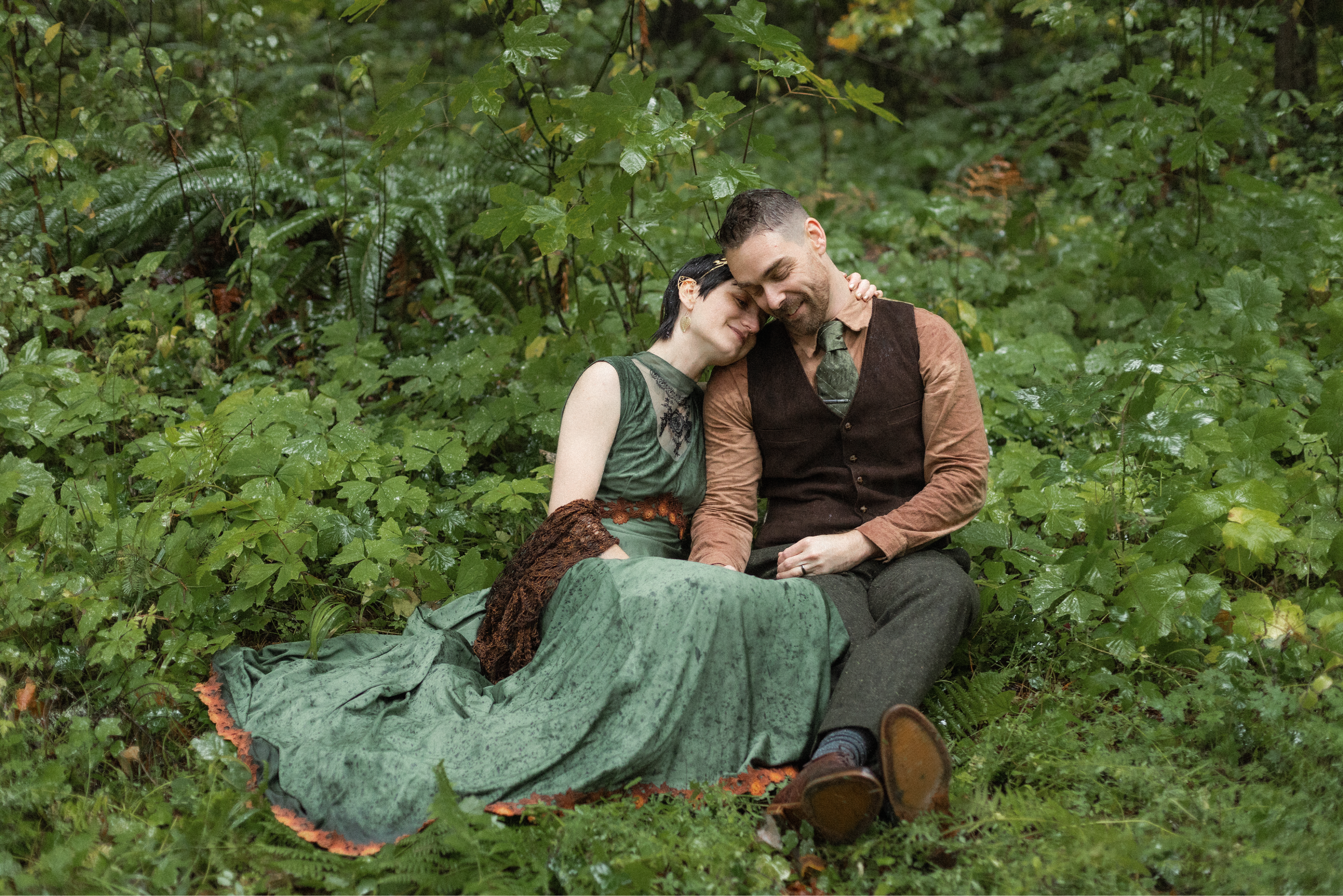 A man and a woman sitting together in a lush green forest, with their heads touching and smiling.