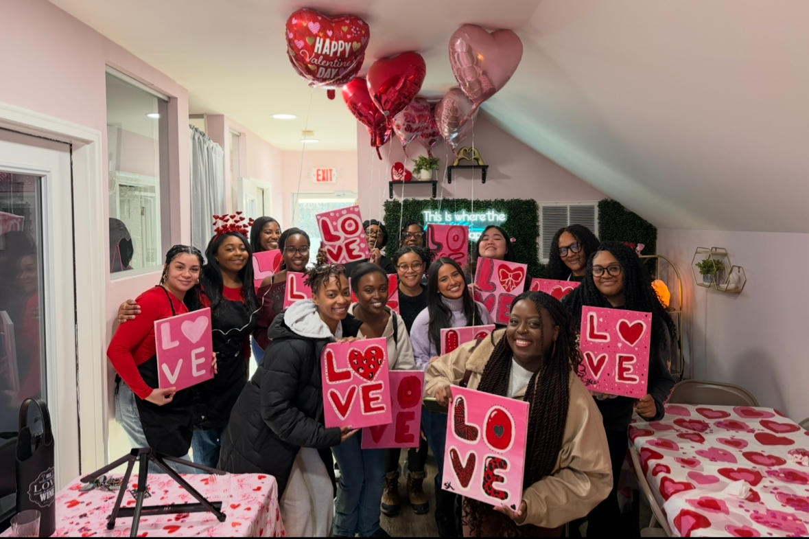 Group of women celebrating Valentine's Day in a decorated indoor space, holding pink signs with 'LOVE' and hearts, with pink and red Valentine's Day balloons overhead.
