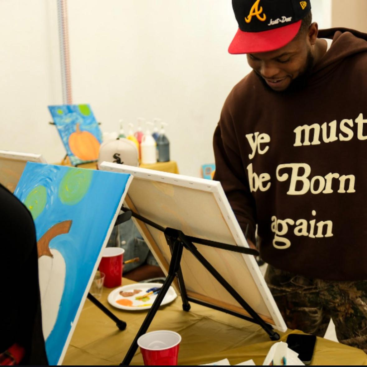 A man wearing a brown hoodie with the phrase "ye must be Born again" and a black and red baseball cap, smiling while painting a colorful canvas at an art event.