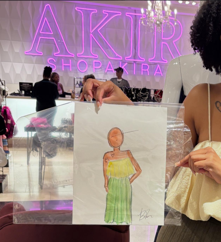 A person holding a plastic bag with a colorful fashion illustration of a woman wearing a yellow top and green skirt, inside a shopping mall decorated with a neon pink sign that reads 'AKIR SHOPAKIRA' in the background.