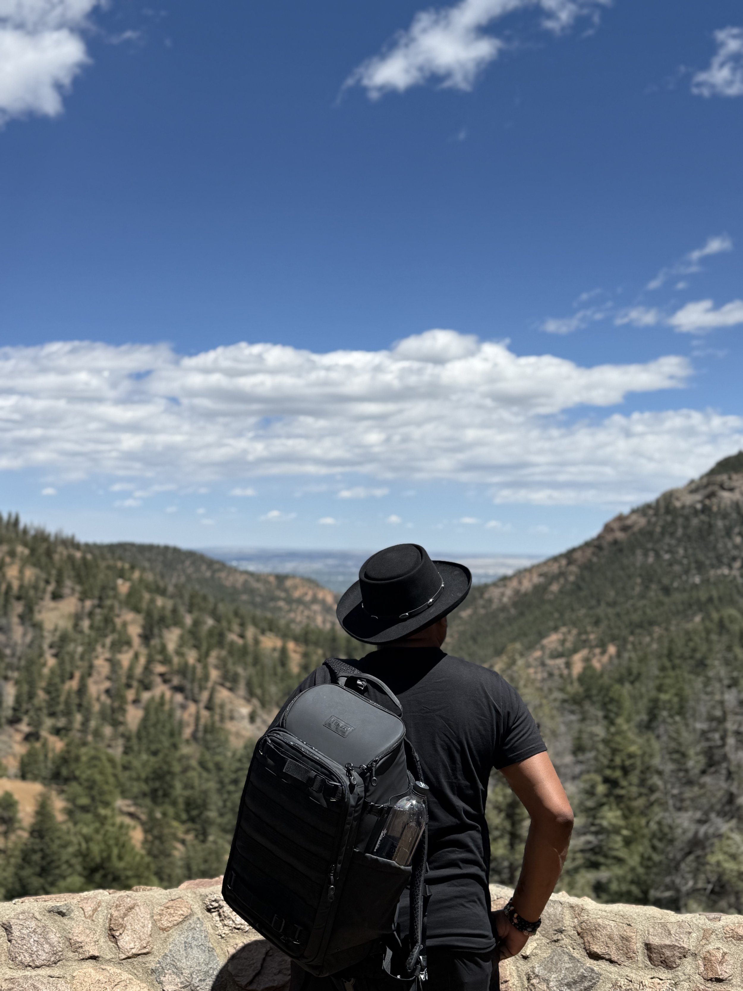 Man with a black hat and black shirt looking at a mountainous landscape with a blue sky and clouds, carrying a backpack with a water bottle.