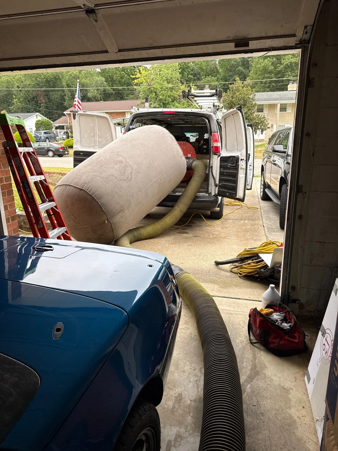 A garage with cleaning equipment and vehicles outside, including a vacuum with a large hose, a blue car, and a white van with open doors.