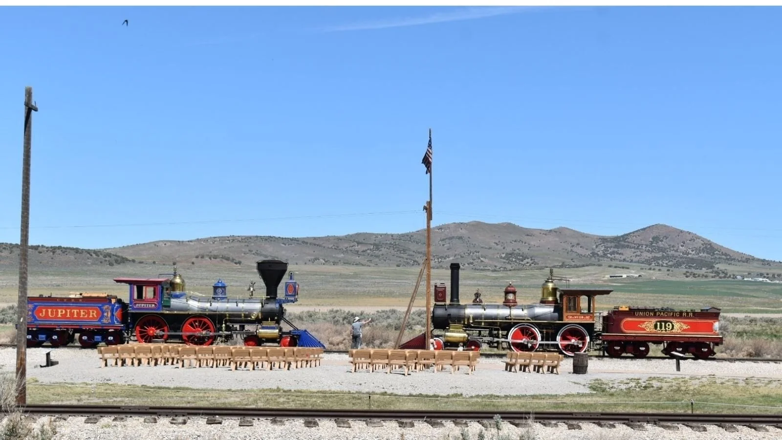 Two vintage steam locomotives displayed outdoors with wooden chairs arranged in front. The left locomotive is labeled 'Jupiter,' and the right one is labeled 'Union Pacific R.R.' A flagpole with an American flag and a pole with a black funnel are bet
