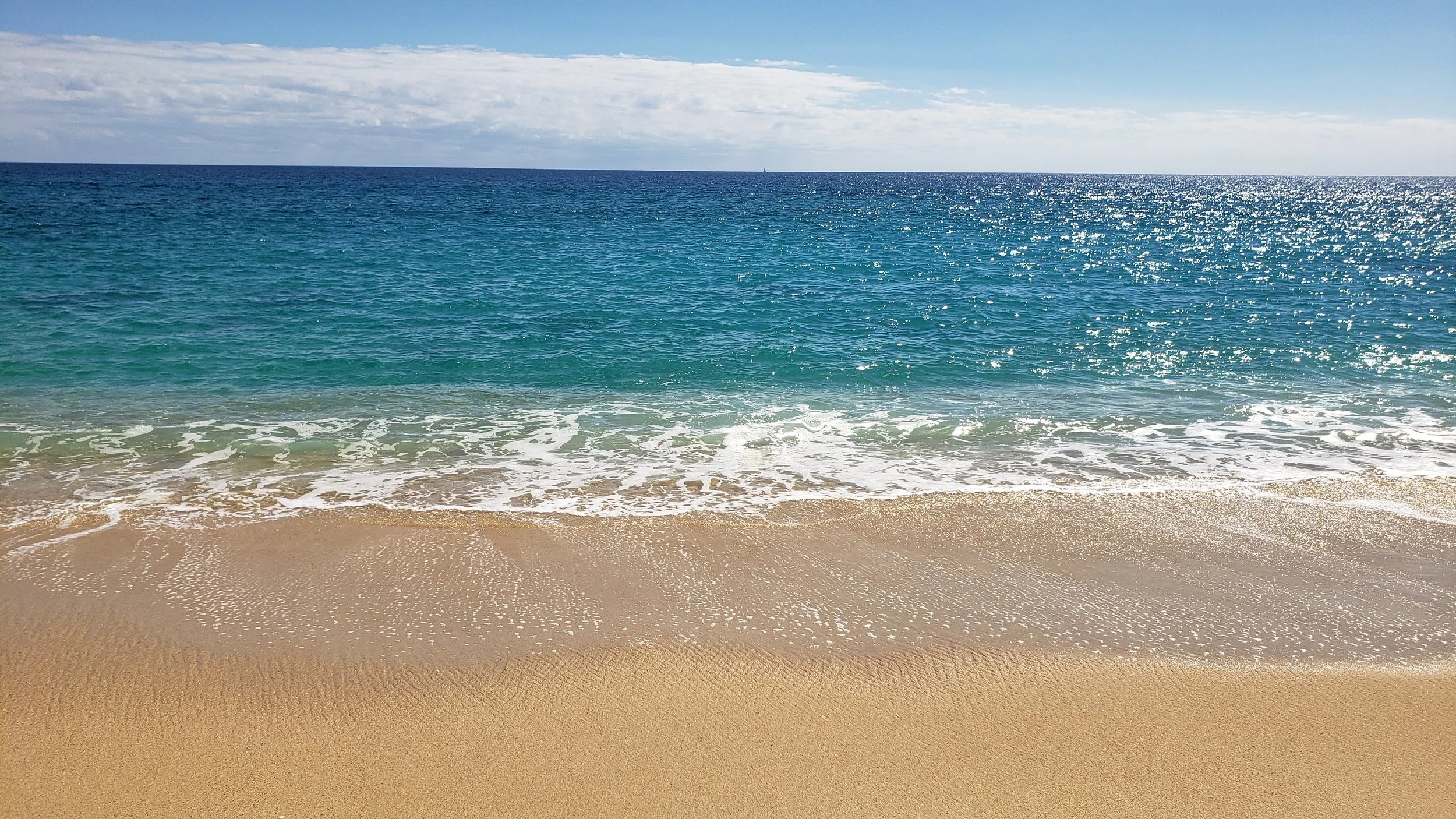 Sunny beach with clear blue ocean waves gently hitting the sandy shore, under a partly cloudy sky.