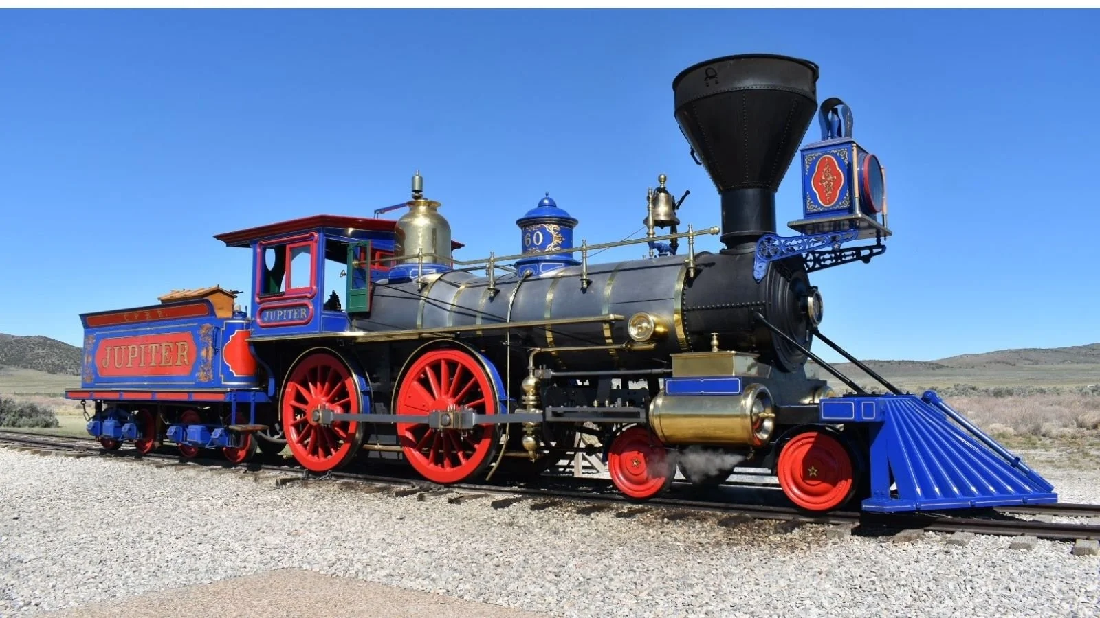 Colorful antique train with red wheels, blue and gold details, and a blue cowcatcher on the front, sitting on train tracks in a rural landscape.