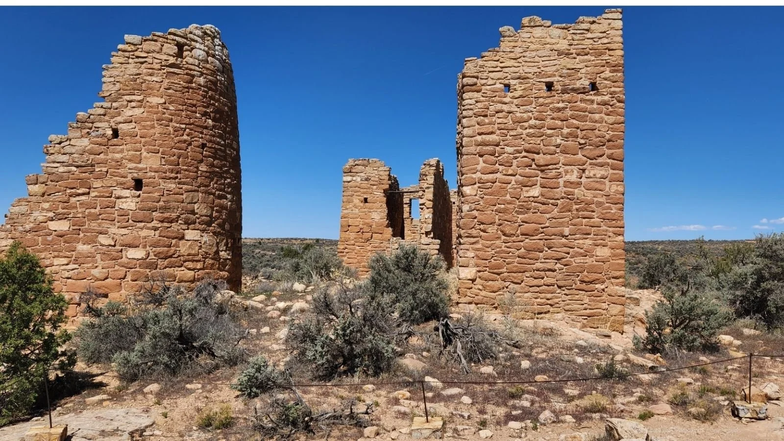Ancient stone ruins of a building in a desert landscape under a clear blue sky with sparse bushes and distant hills.