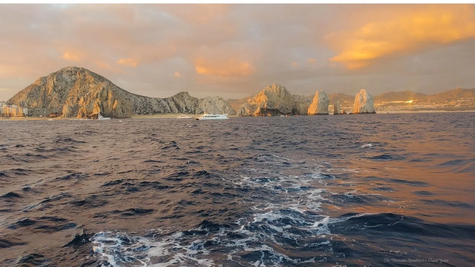 Sea with waves, distant rocky cliffs and formations, boats, and a sunset sky with clouds.