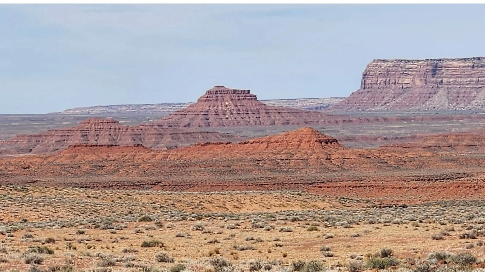 A vast desert landscape with layered red and brown rock formations and mesas under a partly cloudy sky.