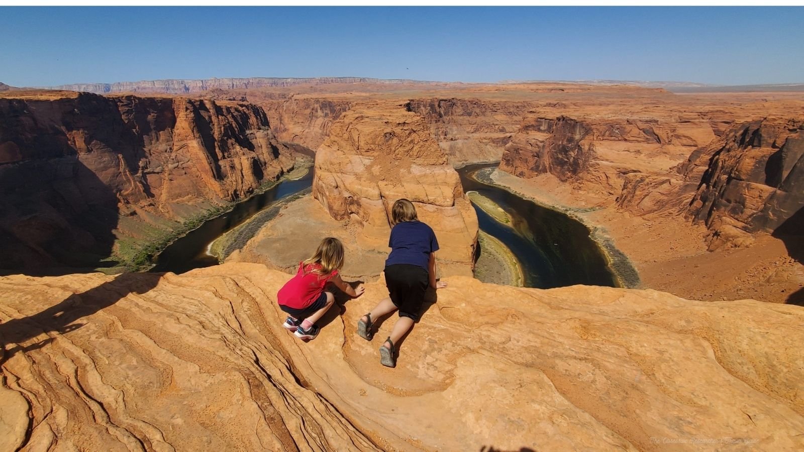 Two children on a rock ledge overlooking the winding Colorado River in Horseshoe Bend, Arizona, during daytime.