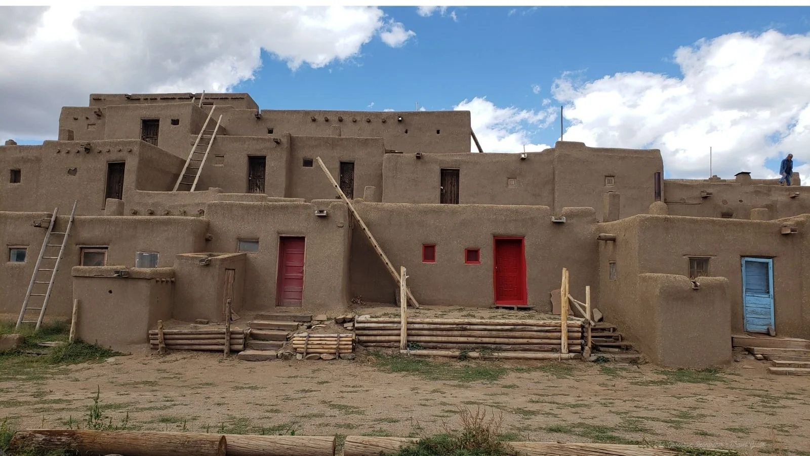 A traditional adobe pueblo with multiple levels, wooden ladders, colorful doors, and steps, against a partly cloudy sky.