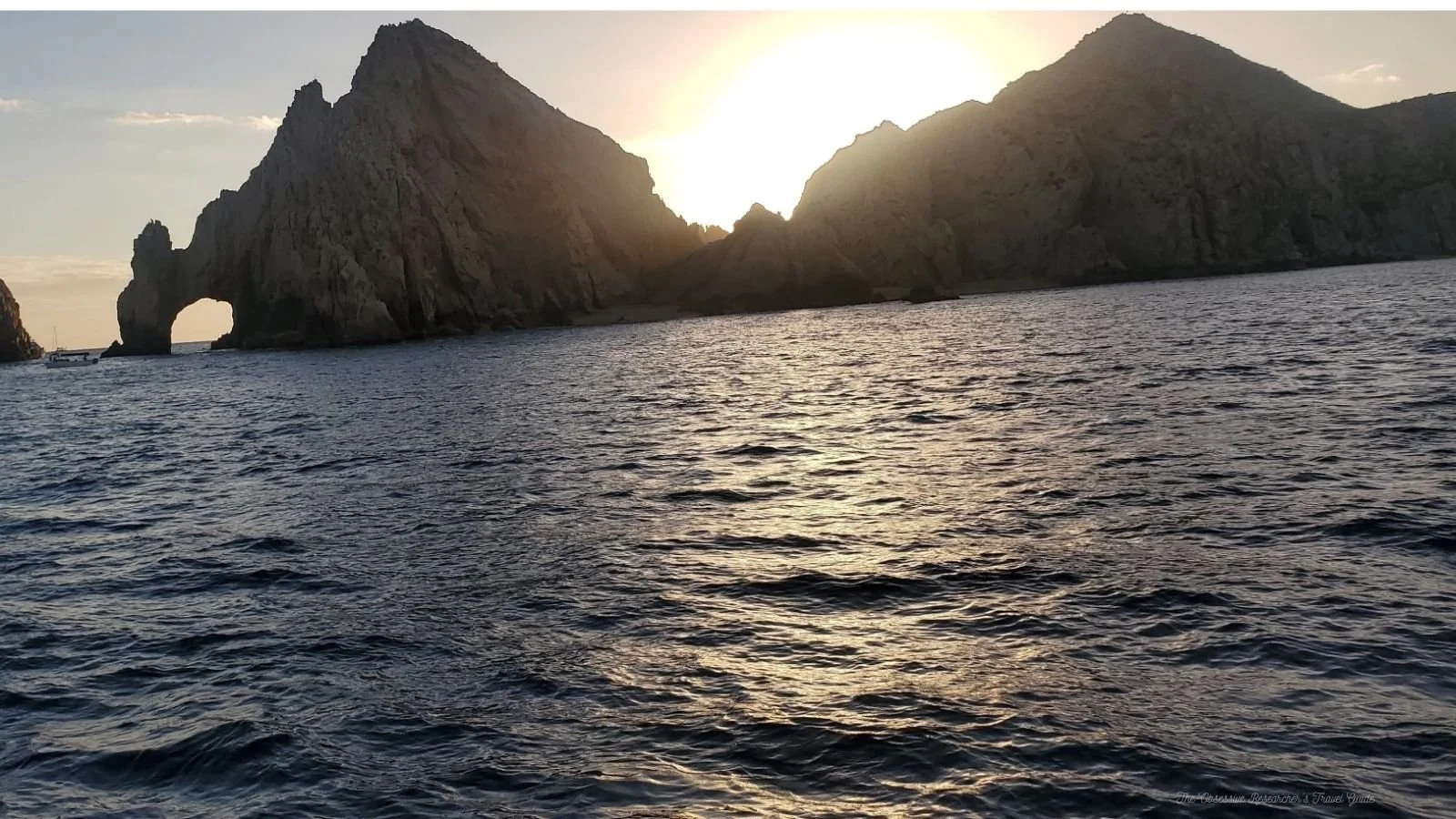 Sea with gentle waves in foreground, large rocky cliffs with an arch and smaller peaks in background, sunset or sunrise lighting.
