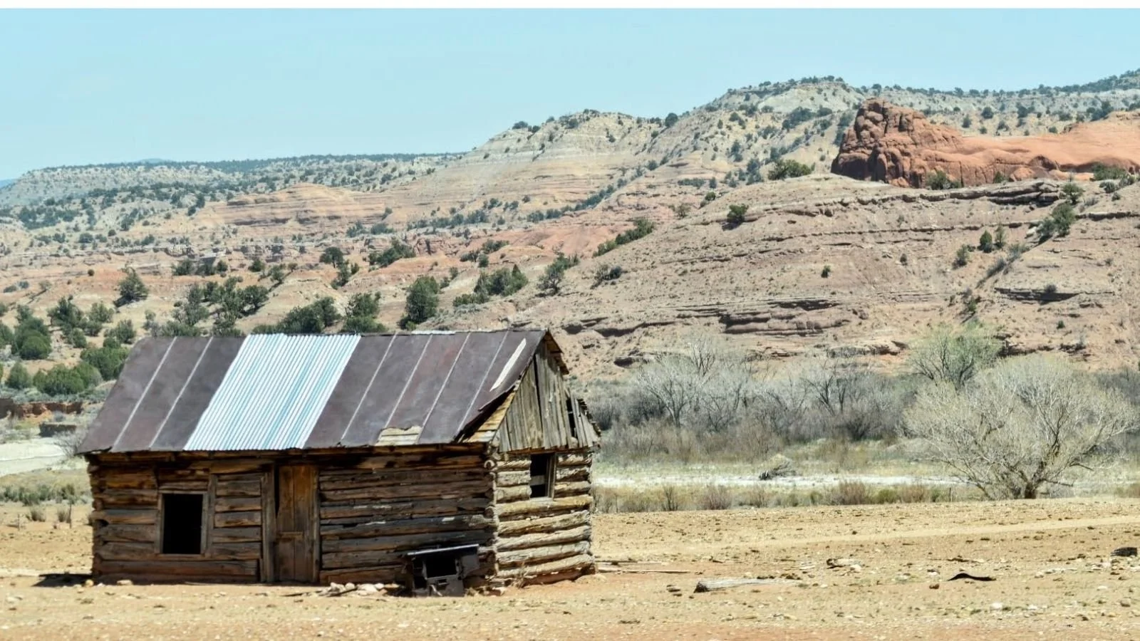 An old wooden cabin with a rusted metal roof in a desert landscape with sparse trees and rocky hills in the background.