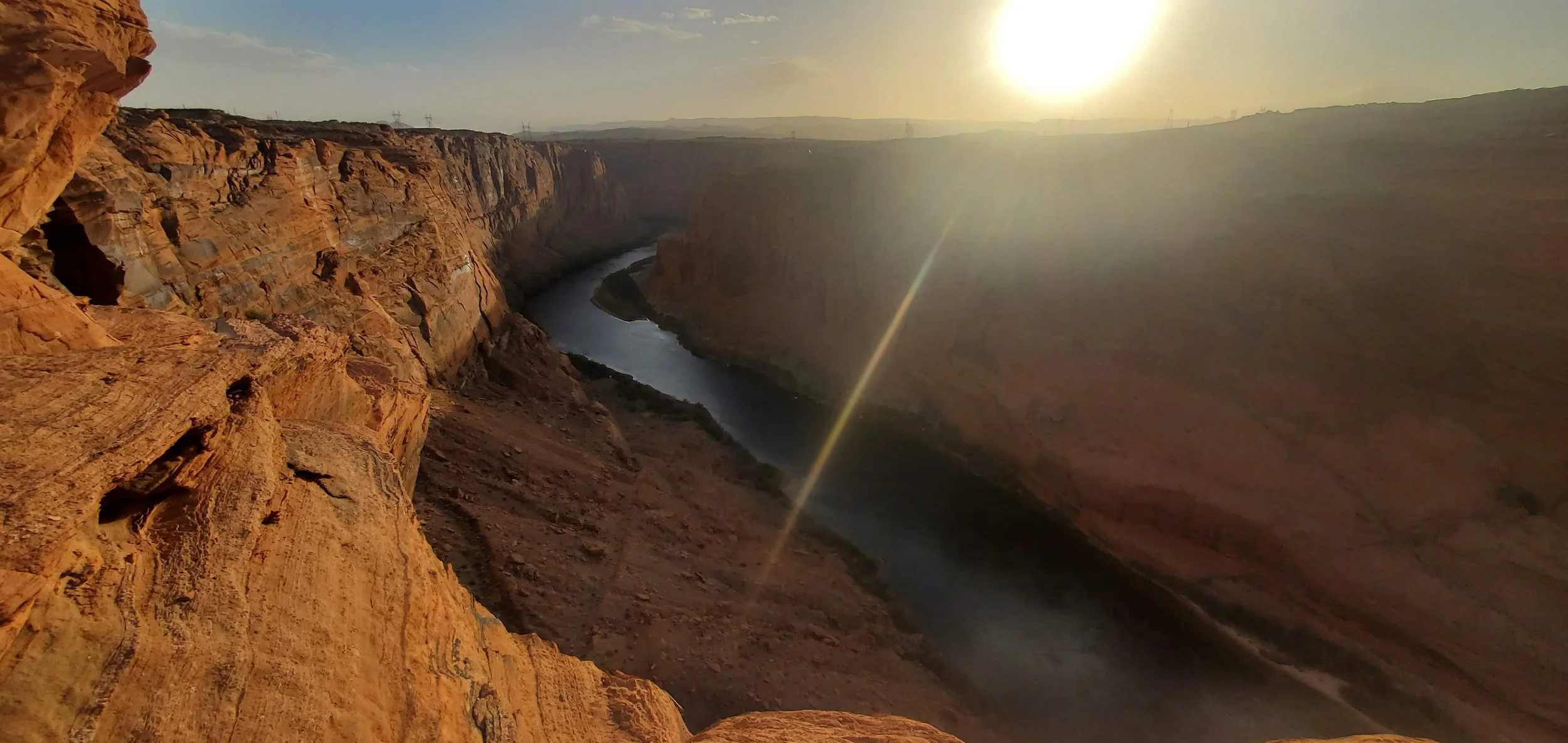 Sunset view over a canyon with a river winding through it, with rugged reddish rock formations on the sides and the sun low in the sky.