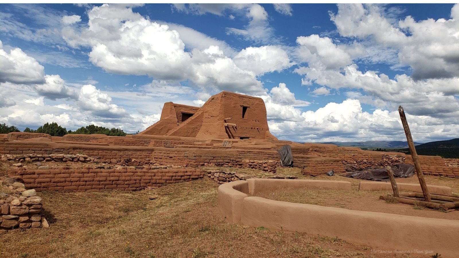 An ancient adobe building with steps leading to it and surrounding brick walls, under a partly cloudy sky.