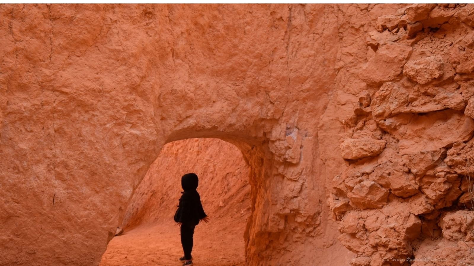 A person standing in a natural sandstone arch formation in a desert landscape with orange rocks and cliffs in the background.