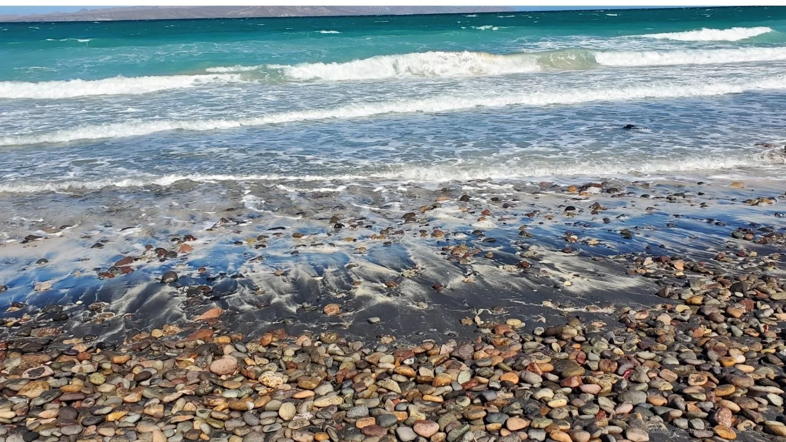 Beach with pebbles in the foreground, gentle waves in the middle, and the ocean extending to the horizon under a clear sky.