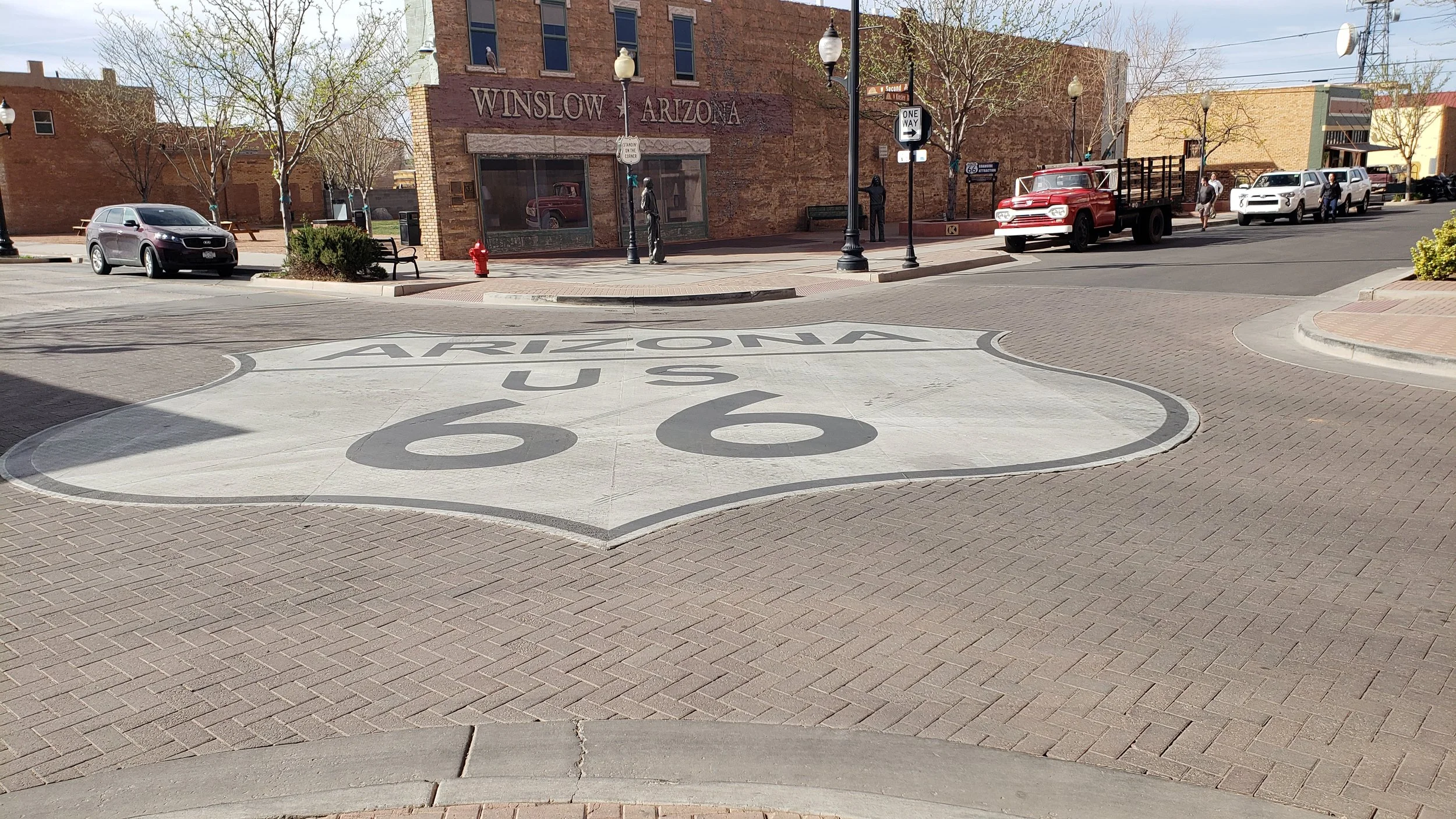 A street scene in Winslow, Arizona, featuring a large Route 66 shield painted on the ground, with vintage and modern vehicles parked along the curb and a brick building with a sign reading 'Winslow Arizona' in the background.