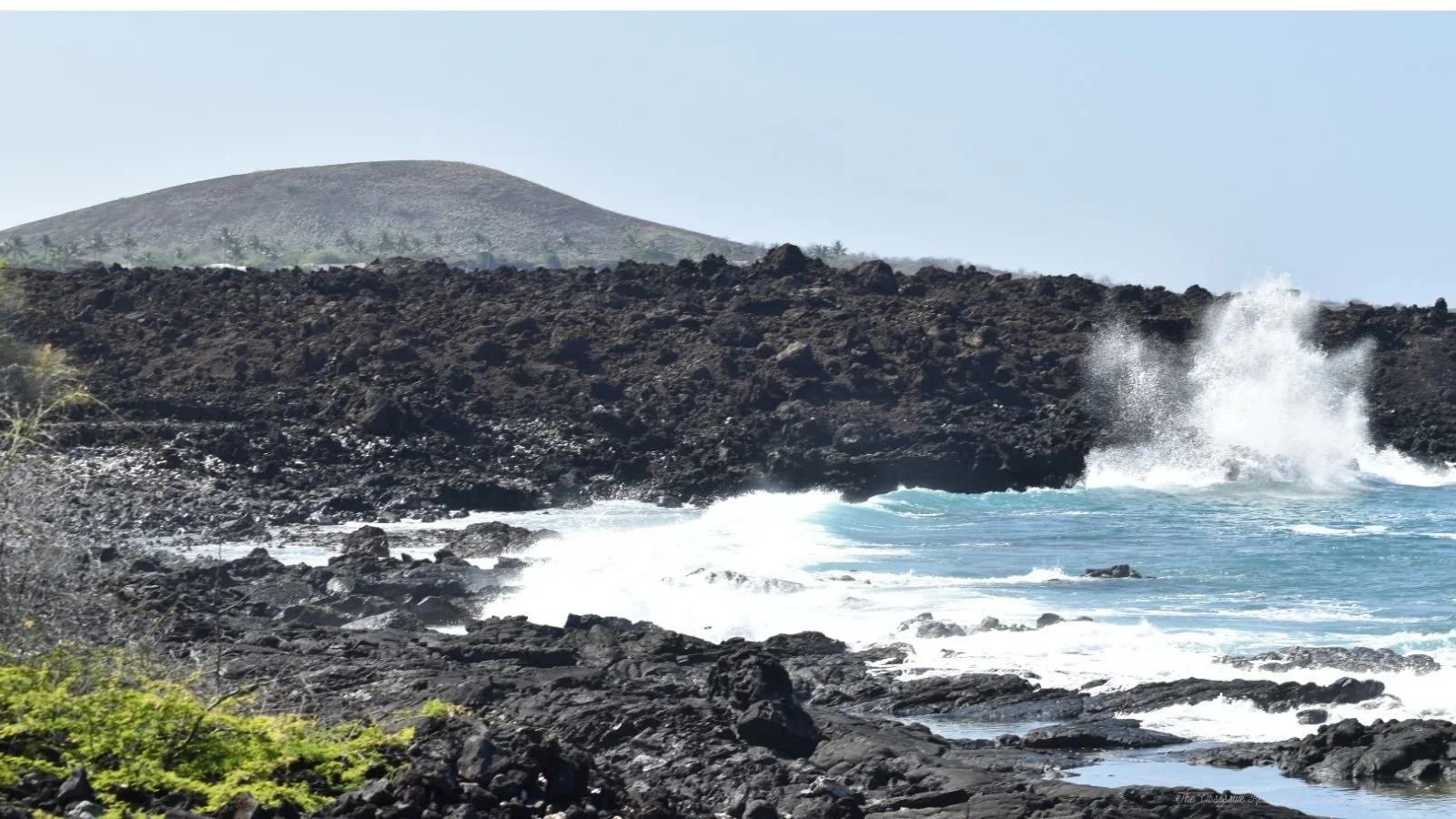 Ocean waves crashing against black volcanic rocks along a coastline with a distant hill in the background.