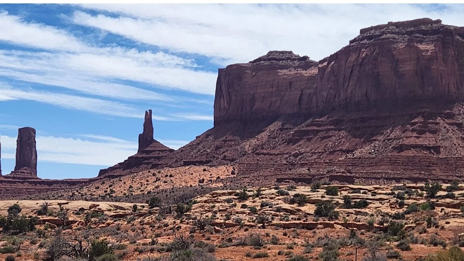 Desert landscape with large red rock formations, mesas, and spires under a partly cloudy sky with sparse desert vegetation in the foreground.