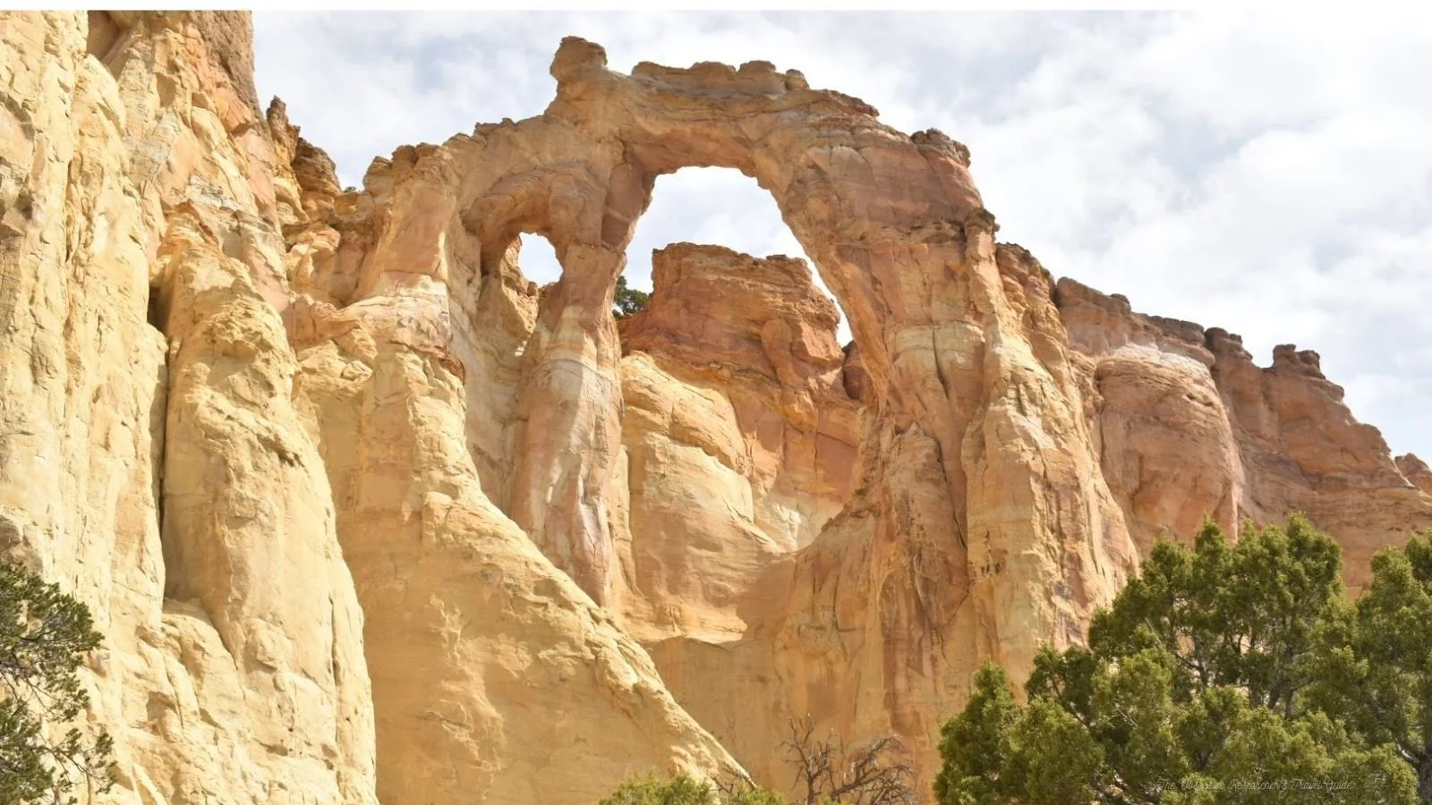 Large sandstone arch formation with eroded openings, surrounded by green trees and cloudy sky.