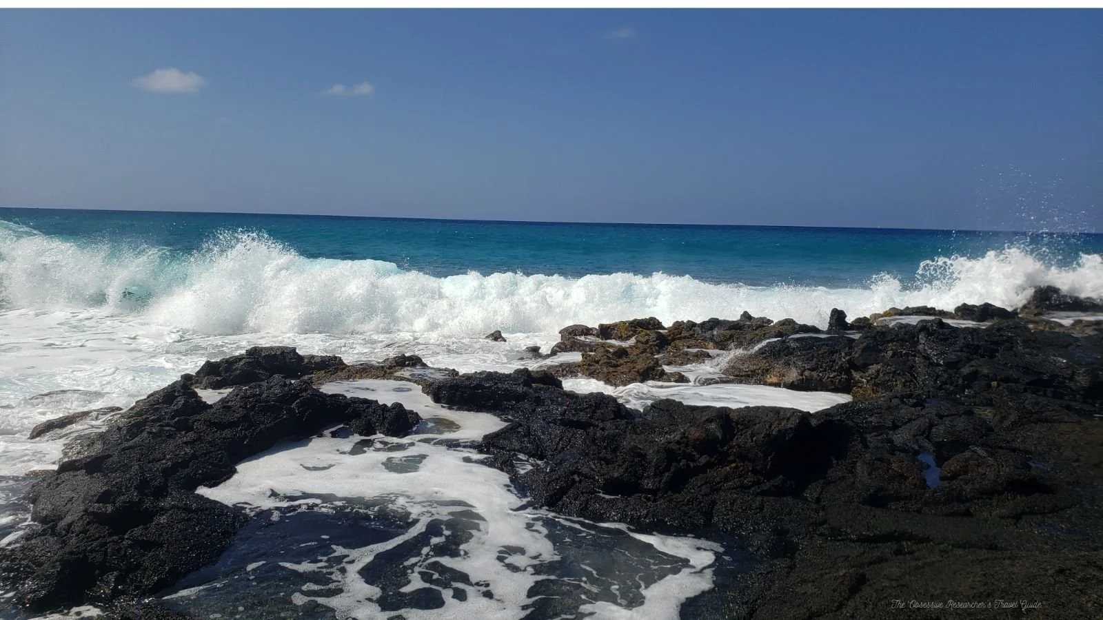 Waves crashing against black volcanic rocks on a sunny beach with a clear blue sky.