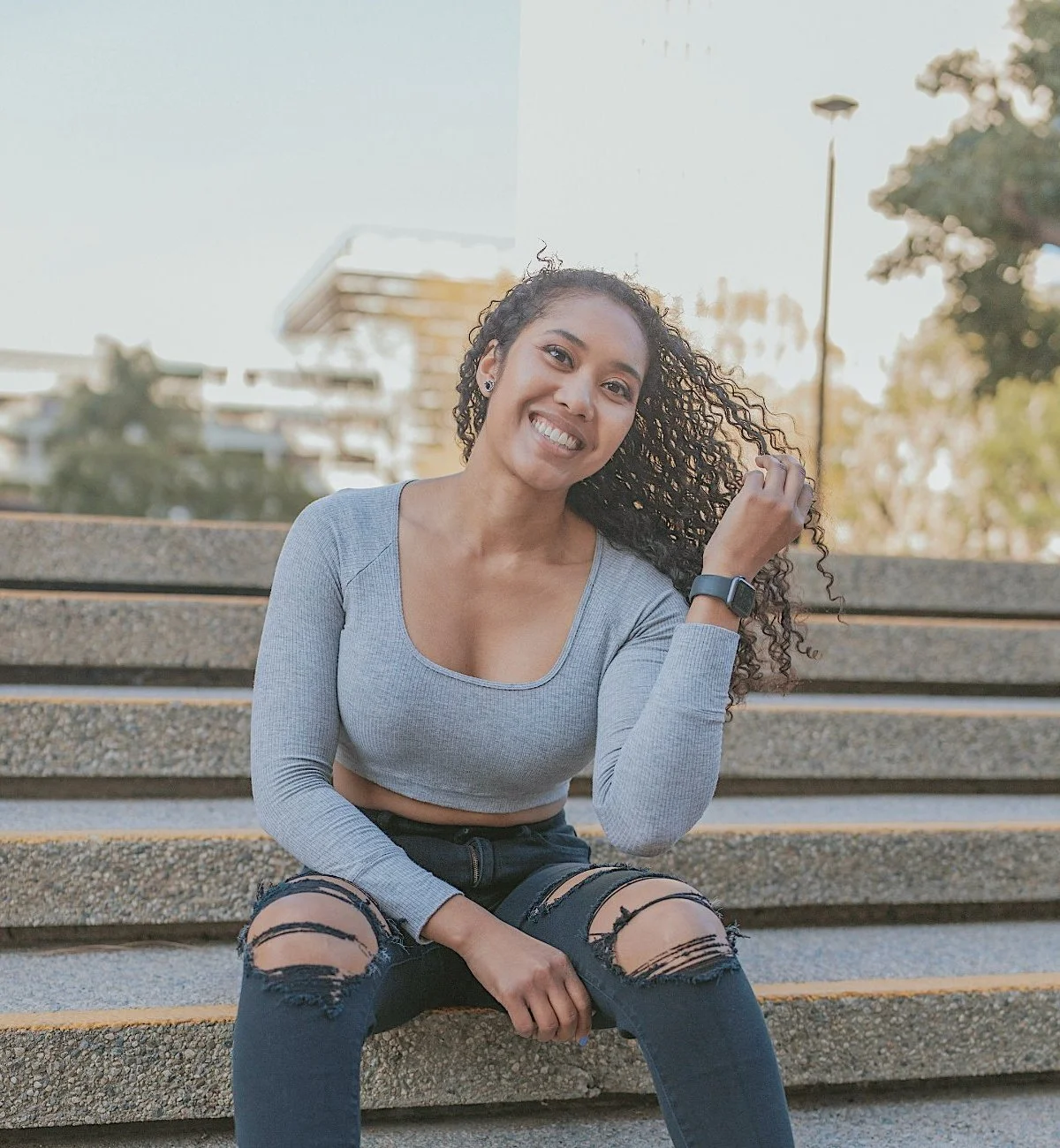 Young woman with curly hair smiling and sitting on outdoor concrete stairs, wearing a gray crop top, ripped jeans, and a smartwatch.