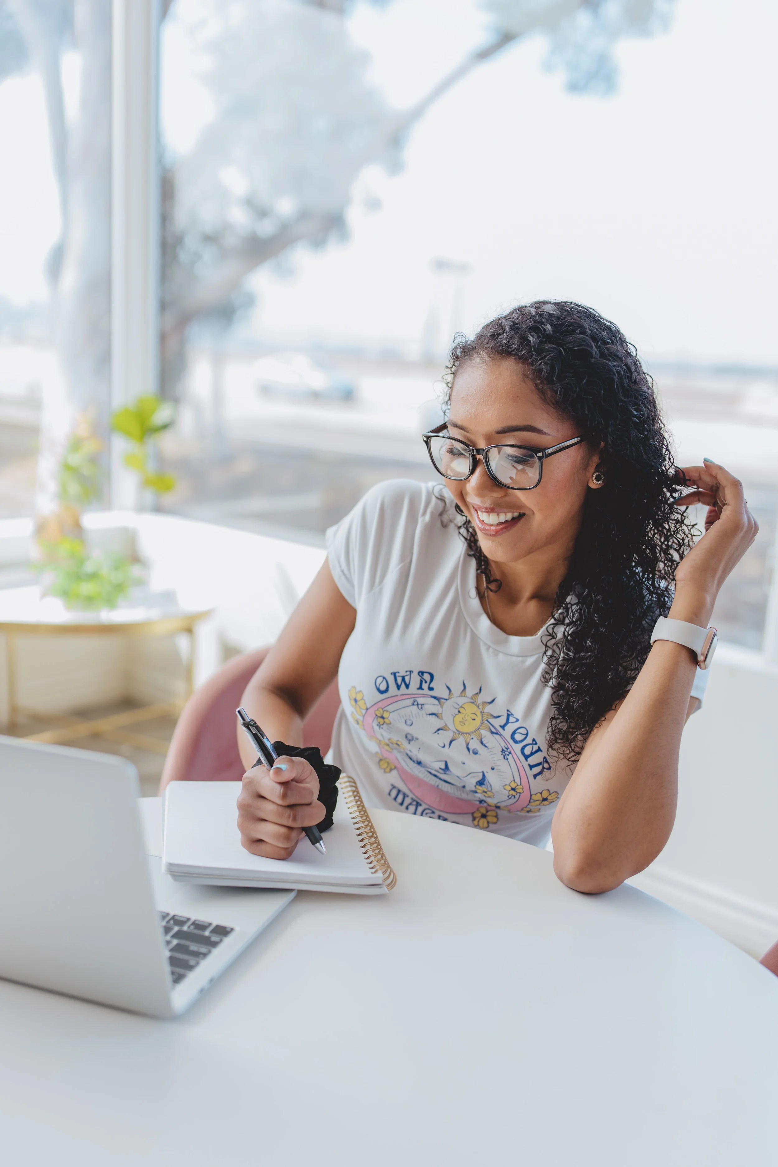 A woman with glasses and curly hair sitting at a white table with a laptop, notebook, and pen, smiling, near large windows with outdoor trees visible.