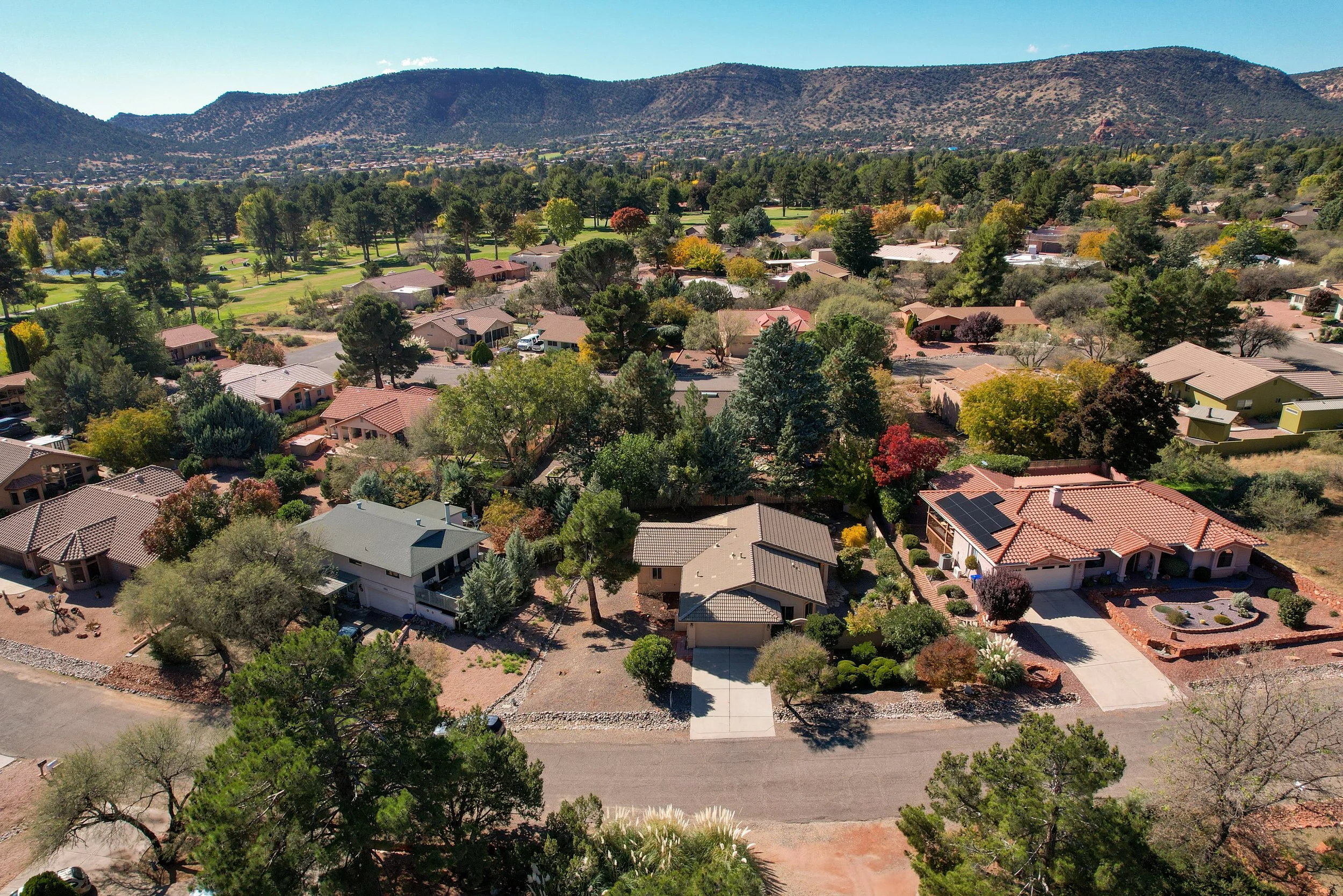 Aerial view of a suburban neighborhood surrounded by trees, with mountains in the background. Houses are situated along a winding street, with well-maintained yards and some solar panels on roofs.