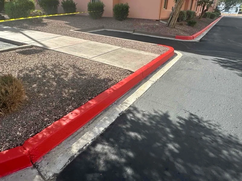 A parking lot curb with a red paint border, sidewalk, and surrounding gravel area with bushes and trees.