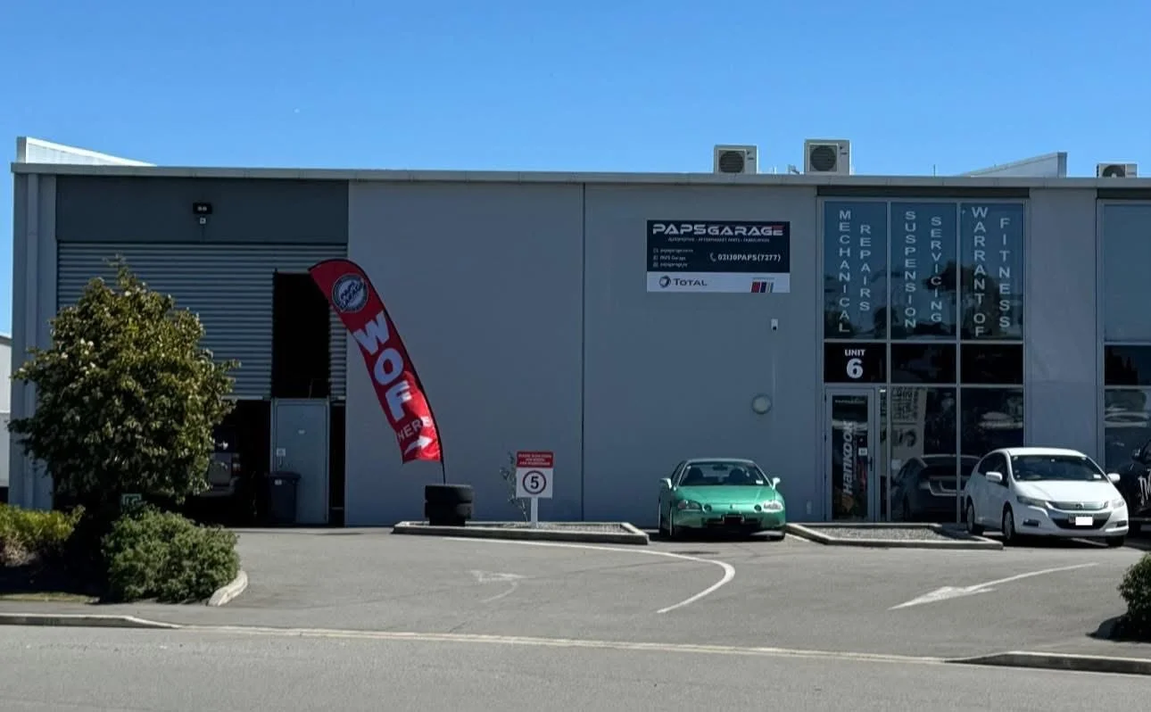 Automotive repair workshop with signage indicating services like mechanical repairs, suspension, servicing, warrant of fitness; multiple cars parked outside, including a green vintage car.