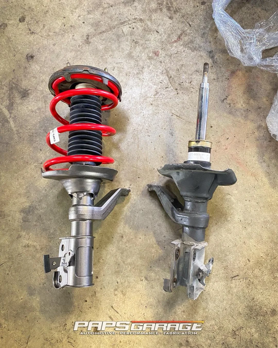 Two car suspension struts laid on a concrete floor, one new with a red coil spring and the other used with a worn appearance.