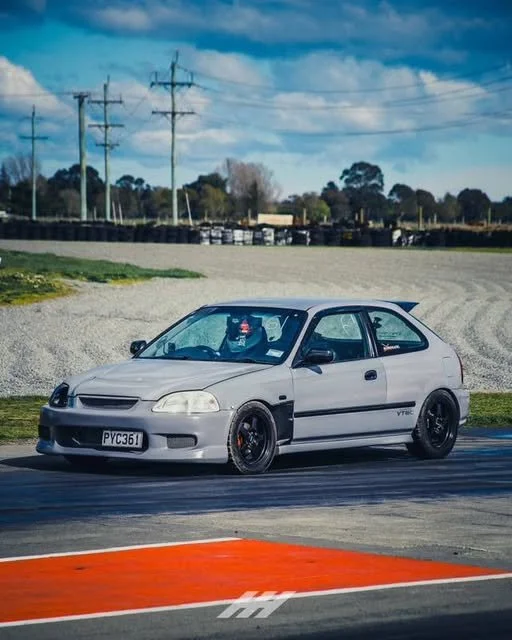 A modified white Honda Civic hatchback on a racetrack with a cloudy sky and power lines in the background.