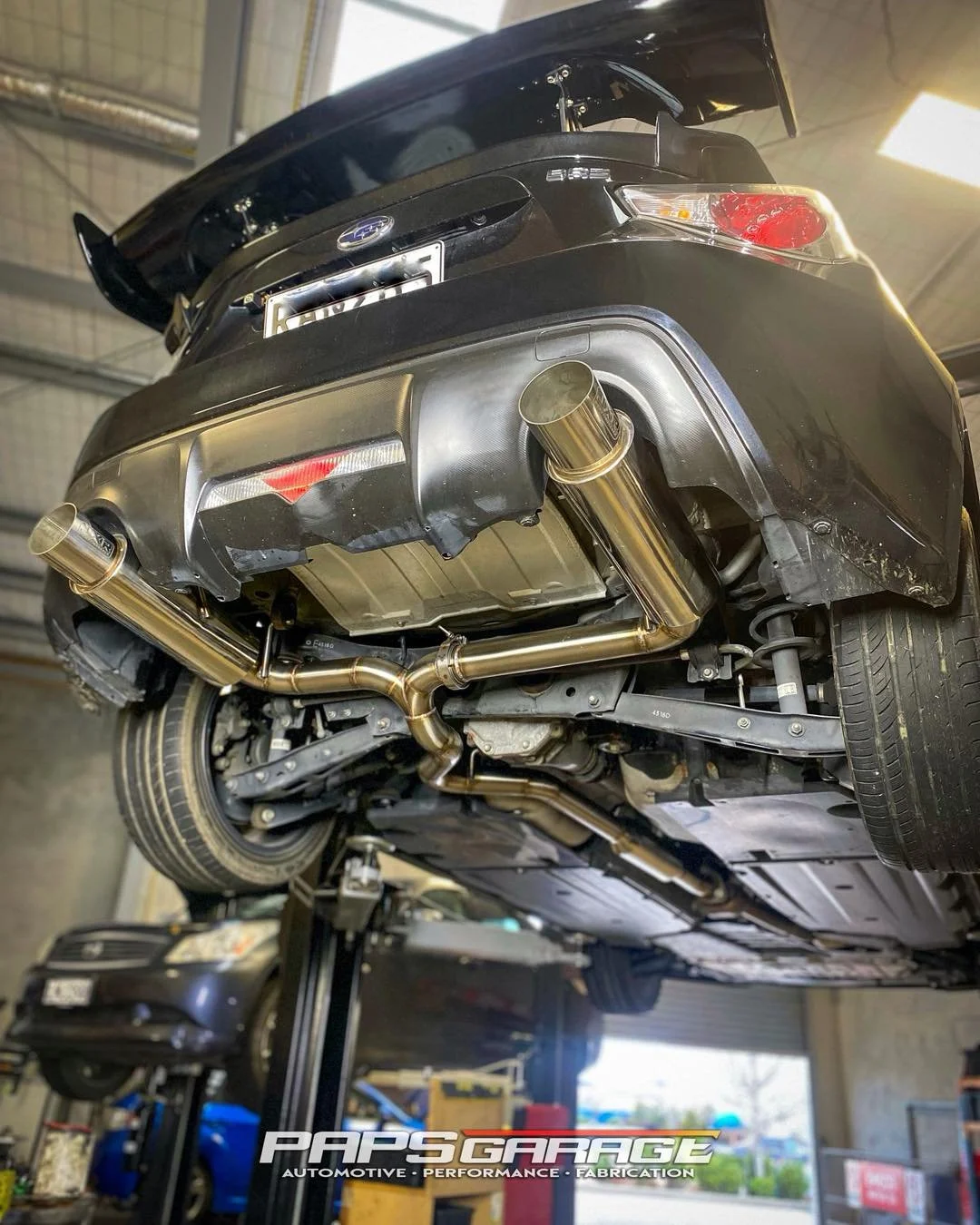 Underside of a black car on a lift showing aftermarket exhaust system with gold-colored pipes.