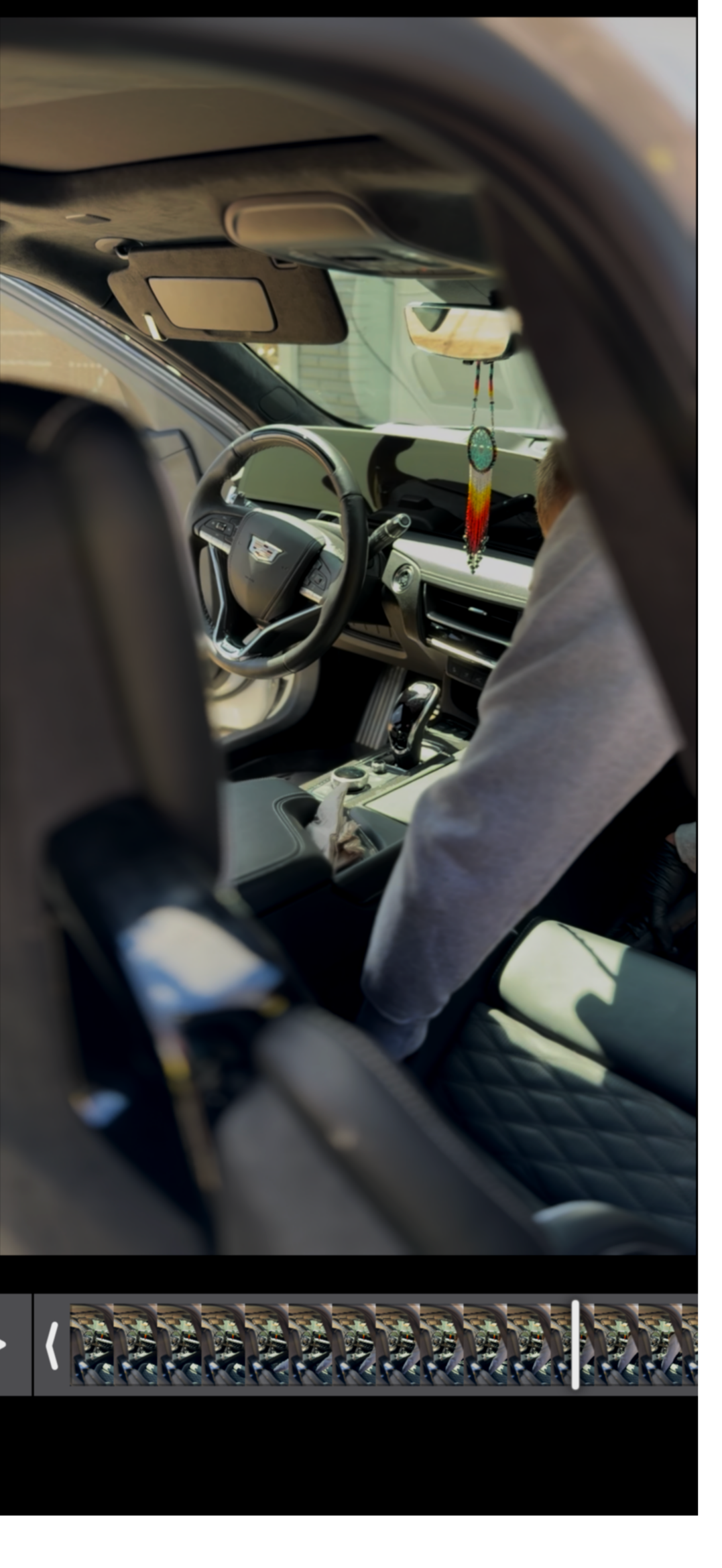 Interior view of a car with a decorative dreamcatcher hanging from the rearview mirror, a steering wheel with a Cadillac logo, and a person in the passenger seat wearing a light-colored hoodie.