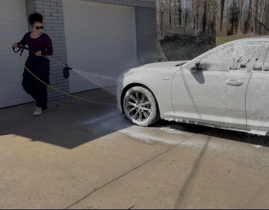 A woman wearing sunglasses and dark clothing is washing a white car with a hose in a driveway outside a house.