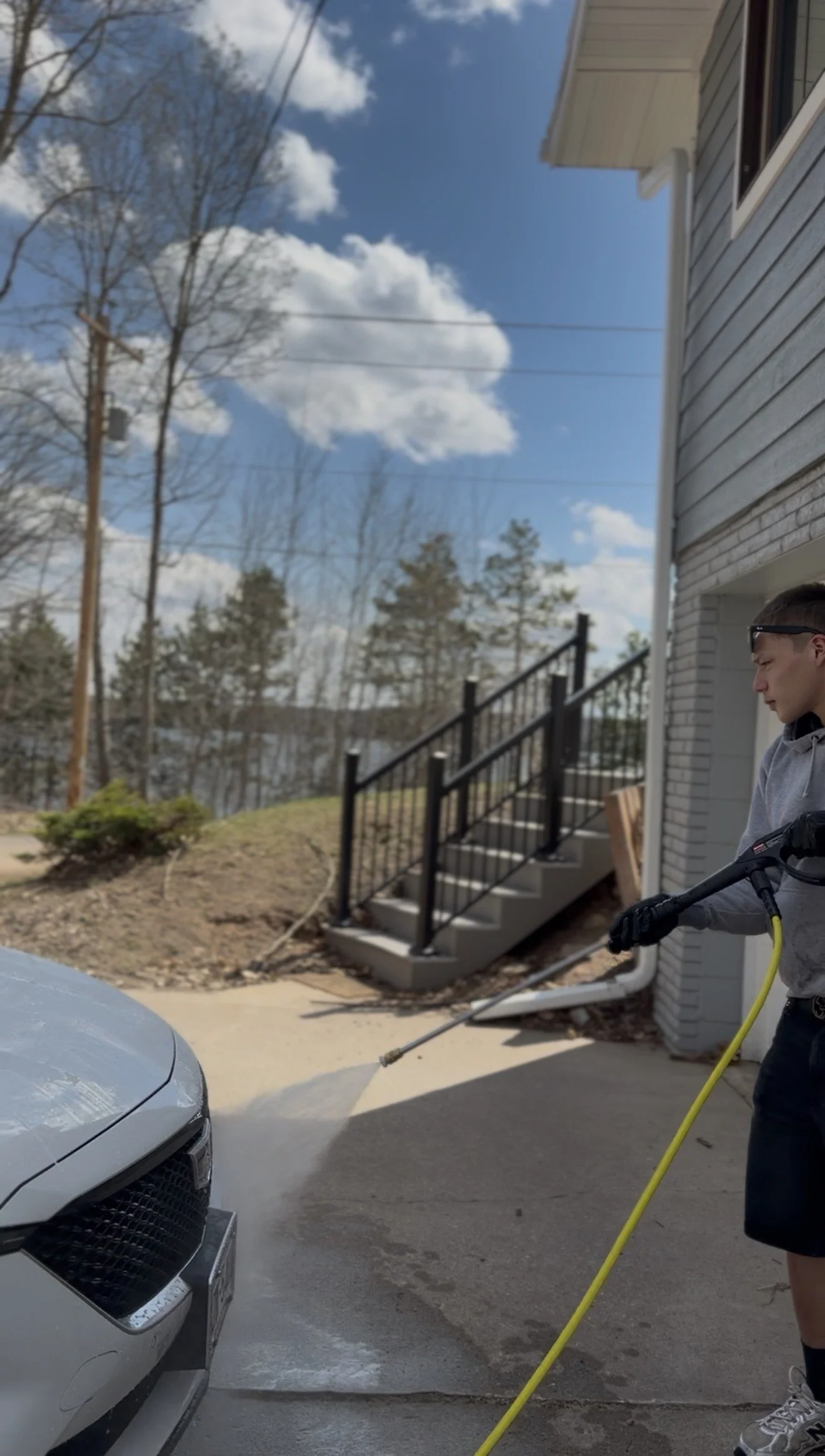 A man wearing glasses, a gray hoodie, and gloves is pressure washing the front of a white car outside a house, with the house's exterior, stairs, and trees in the background.