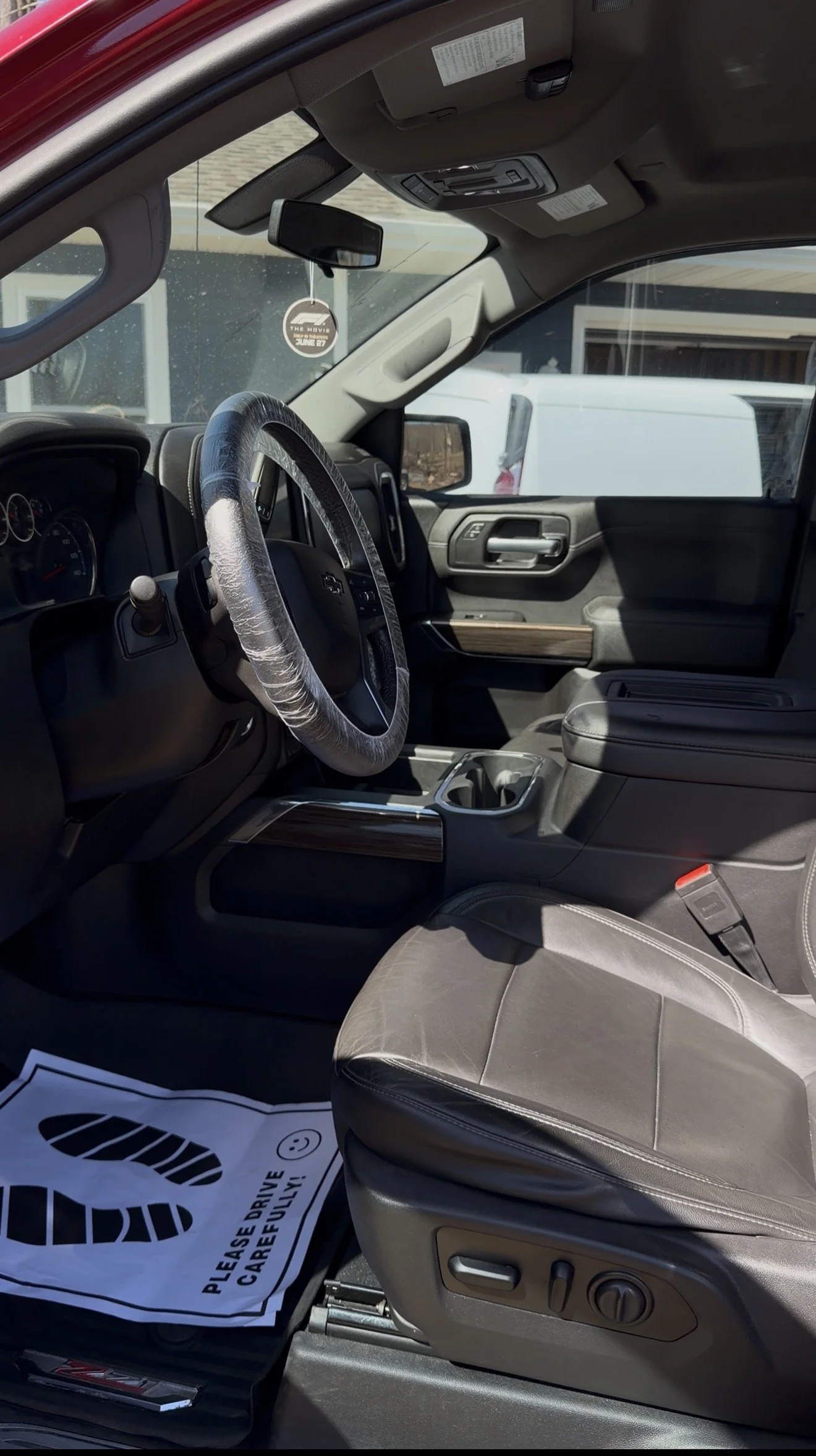 Inside of a vehicle showing the driver's seat, steering wheel with a plastic cover, dashboard with gauges, center console, and a white paper floor mat that says "Please drive carefully!"