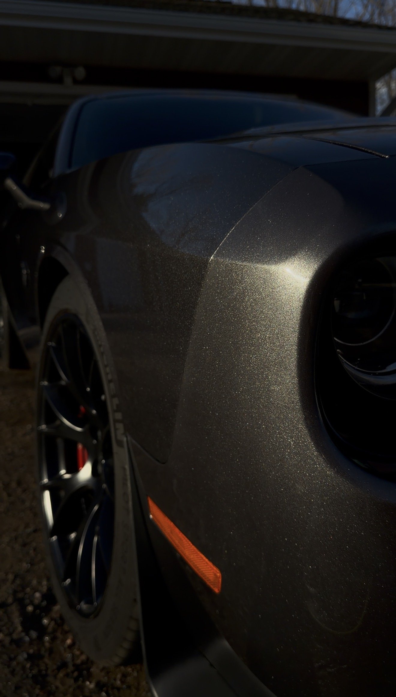 Close-up of a metallic gray sports car with a shiny surface, showing part of the front wheel and vent, parked under a wooden shelter.