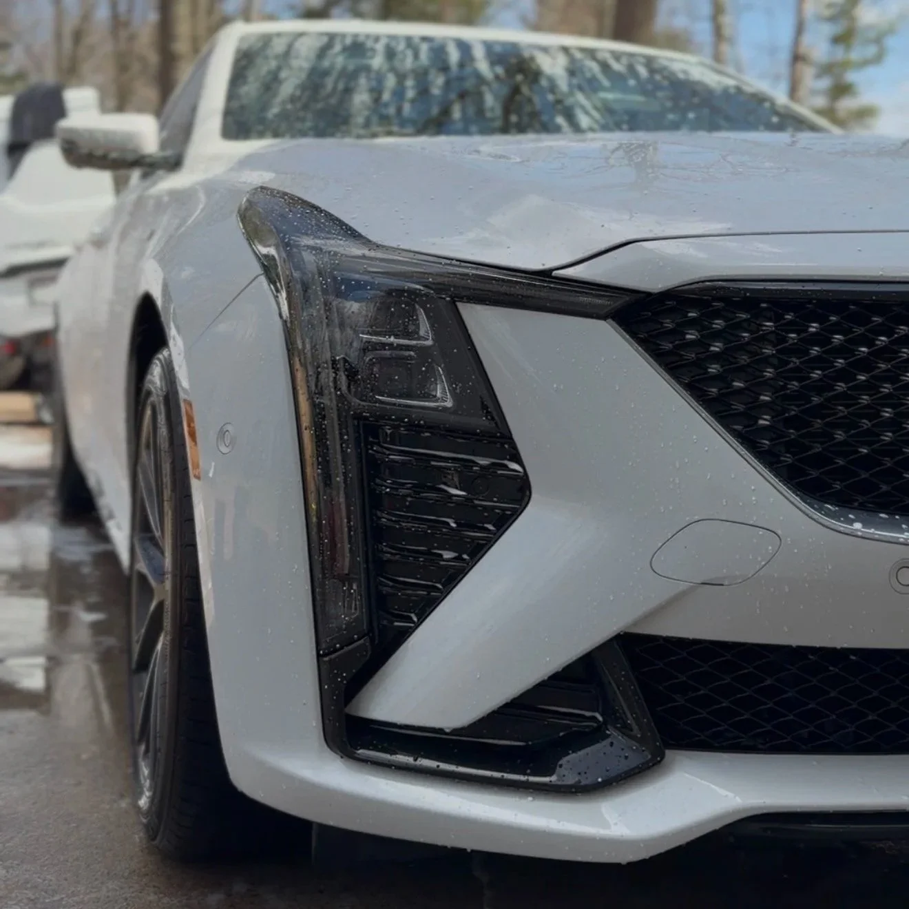 Close-up of the front left side of a modern white luxury car with water droplets on the surface, showing the headlight and grille