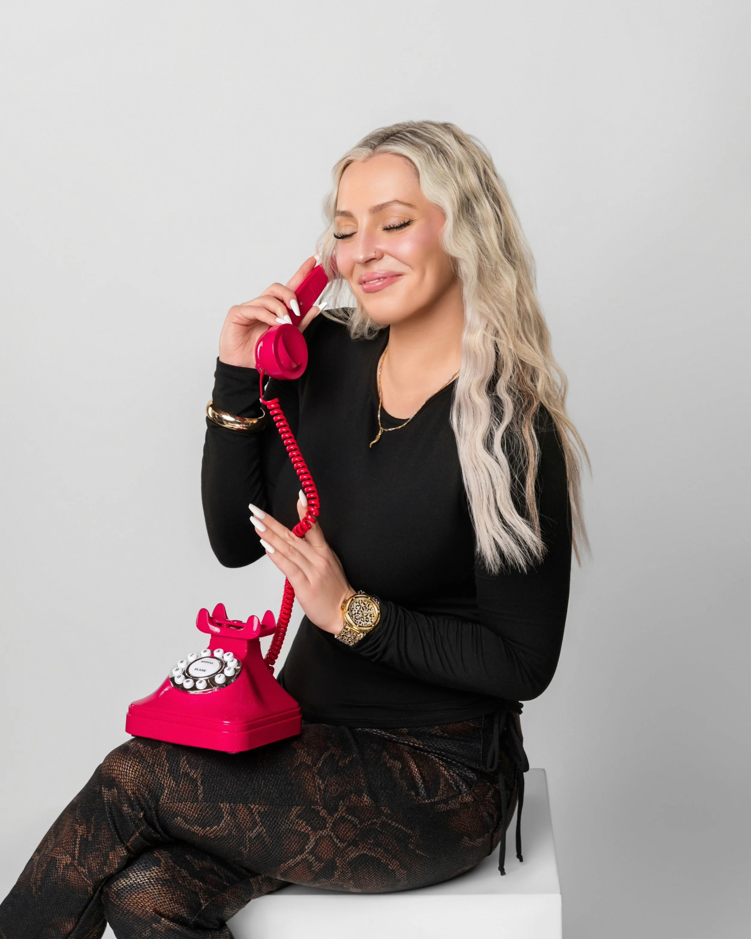 Young woman with blonde hair and a nose piercing, smiling with her eyes closed, holding a pink vintage rotary phone to her ear, sitting in front of a white wall.